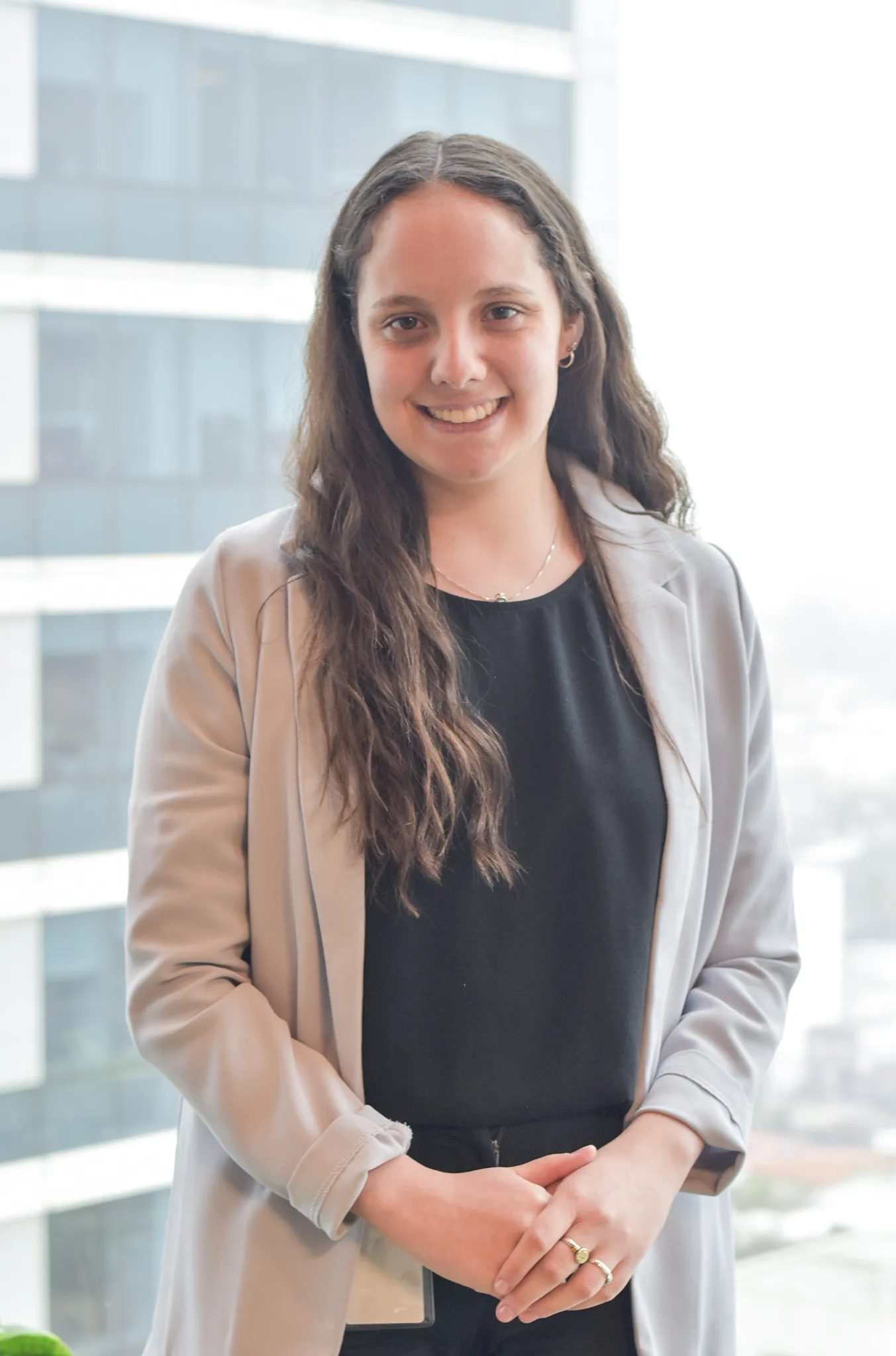 Fotografía profesional de Agustina Villanueva, con cabello largo y rizado, vestida con una blusa negra y un blazer gris claro. Sonríe frente a un fondo de ventanas en un entorno de oficina.