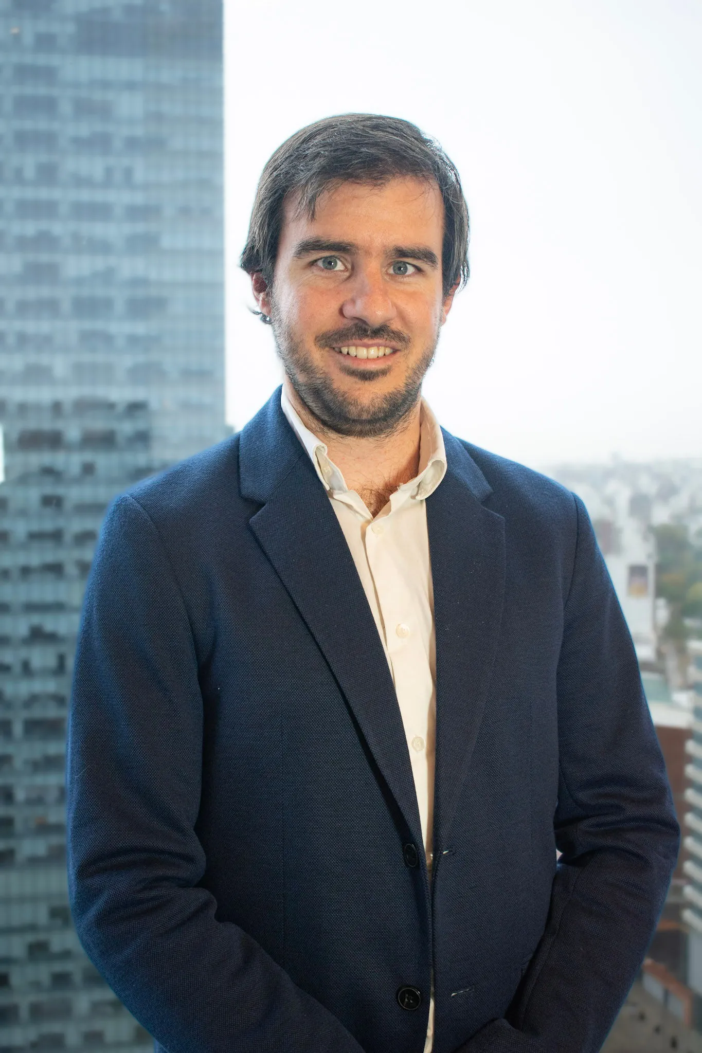 Retrato de Federico Costas, con chaqueta azul y camisa blanca, sonriendo frente a una ventana con vista a la ciudad. Detrás se observa un paisaje urbano moderno con edificios altos.