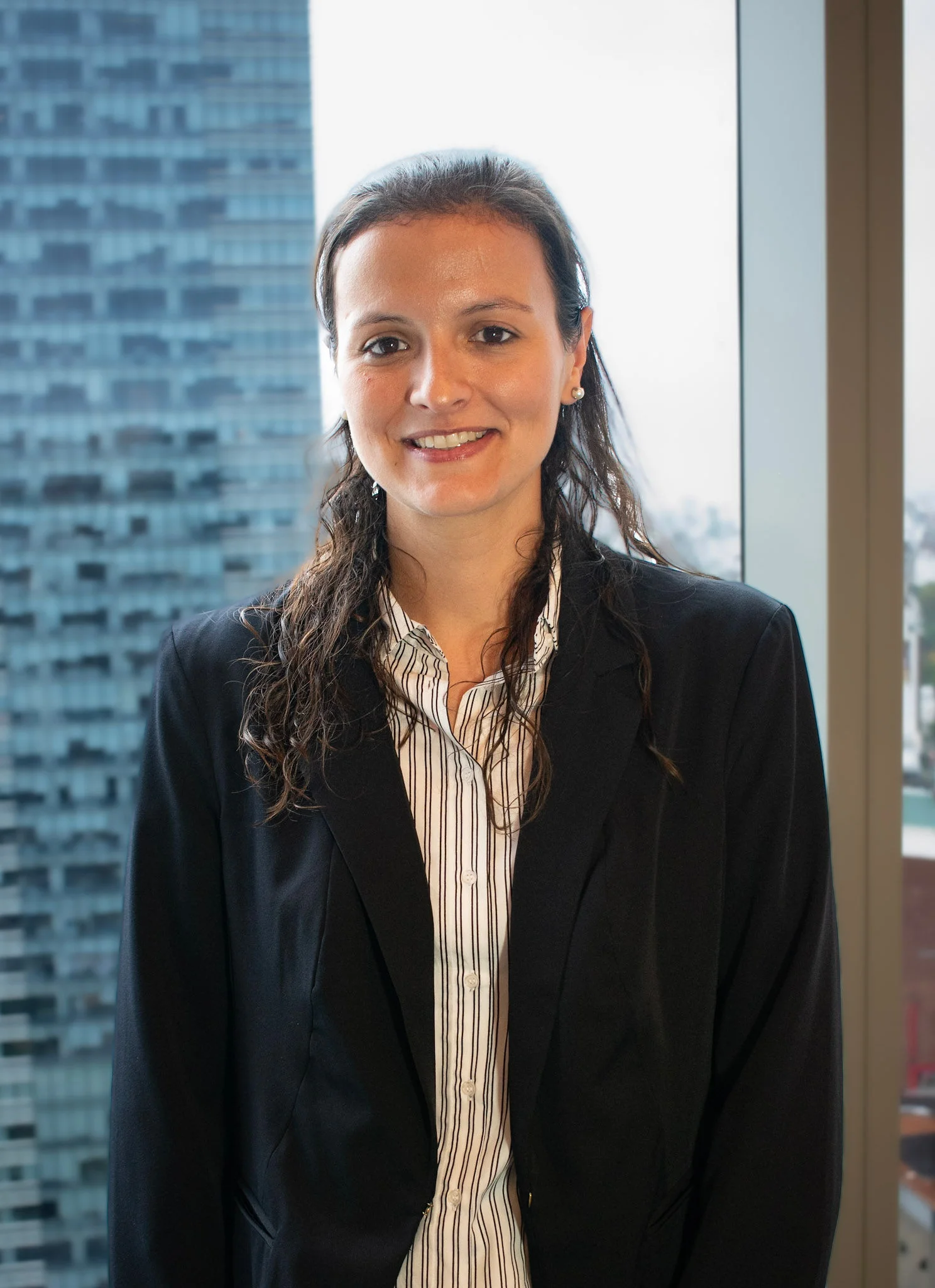 Retrato profesional de Paula Bazzano, contadora en InvestaTrust, posando frente a una ventana con vista a un edificio moderno en una ciudad.
