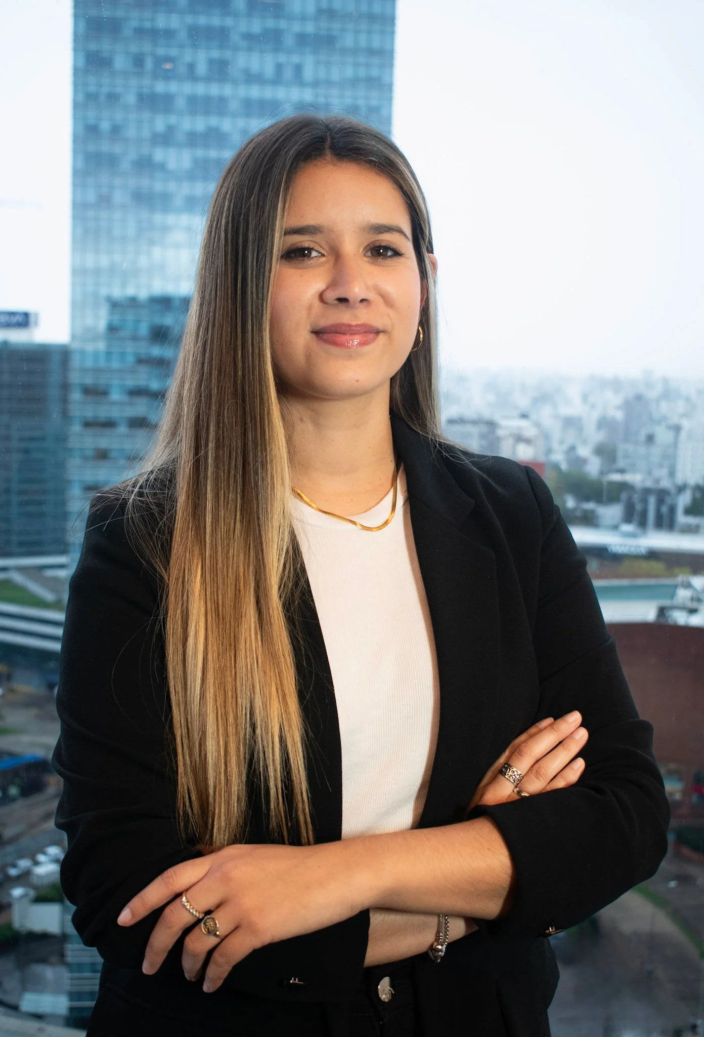 Retrato profesional de Valentina Irigoyen, asistente legal en InvestaTrust, posando con los brazos cruzados frente a un ventanal con vista a un edificio de oficinas.
